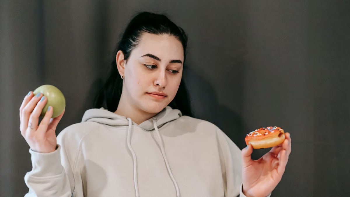 Contemplative plump female in hoodie choosing between ripe green apple and delicious donut in studio